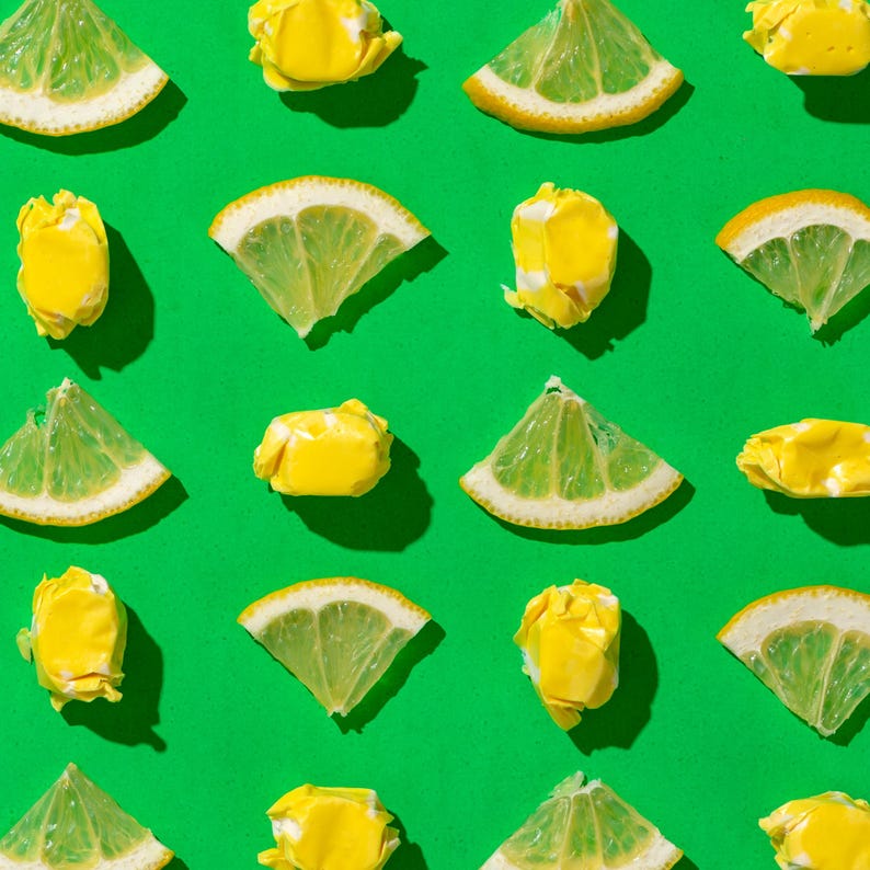 K&ouml;nnte beinhalten: Eine flache Anordnung von gelben Zitronenscheiben und gelben Bonbons auf gr&uuml;nem Hintergrund. Die Zitronenscheiben sind in einem Muster angeordnet, wobei die Bonbons zwischen ihnen verstreut sind.
