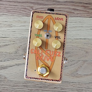 May include: A rectangular guitar effects pedal with a gold faceplate featuring the text CALI FUZZ DRIVE. It includes four gold knobs, a central toggle switch labeled MID CUT, and a large gold footswitch. The border has a brown speckled pattern.