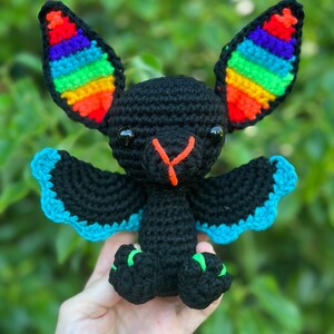 May include: A handmade black crochet bat with rainbow-striped ears, blue wing edges, and green accents on its feet. The bat has orange facial details and black eyes. It is a whimsical, soft toy.