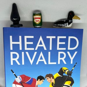 May include: A book titled "HEATED RIVALRY" with a blue cover, featuring hockey players. On top are miniature figurines: a black object, a green "Canada Dry" can, and a painted loon bird.