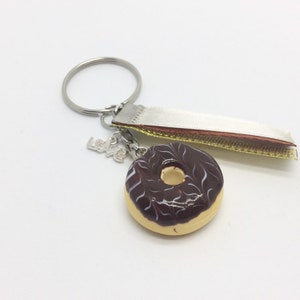 May include: A chocolate glazed donut keychain with white icing and a silver charm that says "Love". The keychain also has a white ribbon with gold trim.