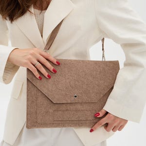 May include: A brown felt laptop bag with a flap closure and a shoulder strap. The bag has a simple, minimalist design and a small pocket on the front. The bag is held by a person wearing a white blazer and pants.