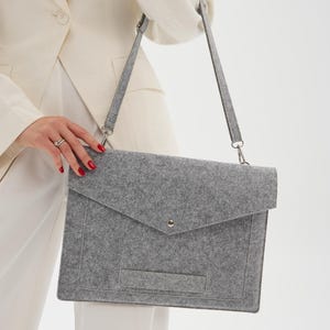 May include: A gray felt messenger bag with a flap closure and a shoulder strap. The bag has a small pocket on the front and a metal clasp. The bag is held by a person wearing a white blazer and pants.