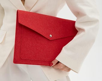 Könnte beinhalten: Eine leuchtend rote Laptop-Hülle aus Filz mit Klappe und silbernem Druckknopf. Die Hülle hat eine Vordertasche und wird von einer Person in einem weißen Blazer und Hose gehalten. Das Thema ist Bürobedarf.