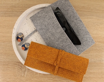 Puede incluir: Una bolsa de fieltro gris con un par de gafas de sol negras en su interior. Una bolsa de fieltro marrón más pequeña está encima de la bolsa gris. Ambas bolsas están sobre una superficie moteada blanca y gris.