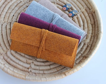 Puede incluir: Cuatro bolsas de fieltro de diferentes colores, apiladas unas sobre otras. Las bolsas son de color amarillo mostaza, burdeos, gris claro y marrón. Las bolsas están en una cesta tejida.