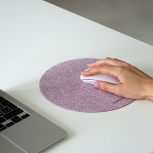Puede incluir: Una alfombrilla de ratón redonda de fieltro morado con la palabra "QUDZYY" impresa en letras doradas. Un ratón inalámbrico blanco está sobre la alfombrilla.