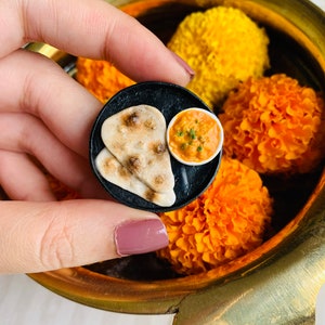 Puede incluir: Un modelo de arcilla en miniatura de un plato con dos panes naan y un pequeño cuenco de curry. El modelo está siendo sostenido por una mano contra un fondo de flores de caléndula naranja.