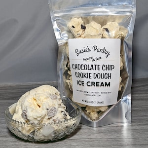 May include: A silver resealable bag filled with freeze-dried chocolate chip cookie dough ice cream. The bag has a white label with the text "Susie's Pantry freeze dried Chocolate Chip Cookie Dough Ice Cream" and the company's contact information. The bag is sitting on a wooden surface next to a glass bowl filled with vanilla ice cream.