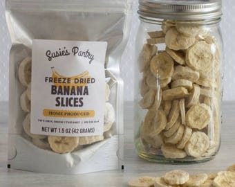 May include: A clear plastic bag filled with freeze-dried banana slices. The bag has a white label that reads "Susies Pantry Freeze Dried Banana Slices Home Produced Net Wt 1.5 oz (42 grams)". A clear glass jar filled with freeze-dried banana slices is next to the bag.
