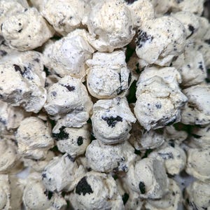 Pu&ograve; includere: Primo piano di un mucchio di dolcetti bianchi di forma irregolare con inclusioni scure e macchiettate. I dolcetti hanno una consistenza leggermente ruvida e sembrano essere un tipo di snack o confetteria, forse al gusto biscotti e panna.