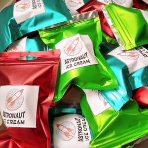 Pu&ograve; includere: Un mucchio di sacchetti di alluminio colorati di gelato per astronauti. I sacchetti sono in tonalit&agrave; di rosso, verde e verde acqua. Ogni sacchetto ha un'etichetta bianca con le parole "Astronaut Ice Cream" e una grafica di razzo.