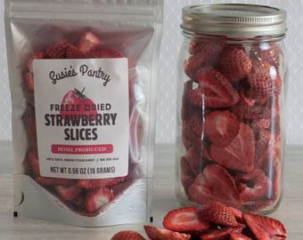 May include: A clear plastic bag filled with freeze-dried strawberry slices. The bag has a label that reads "Susies Pantry Freeze Dried Strawberry Slices Home Produced Net Wt 0.55 oz (15 grams)". A clear glass jar filled with freeze-dried strawberry slices is next to the bag.