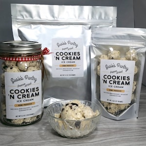 May include: A clear glass jar filled with freeze-dried cookies 'n cream ice cream. The jar has a red and white checkered ribbon around it. The label on the jar reads "Susie's Pantry freeze dried Cookies 'n Cream Ice Cream Home Produced 348 E 320 N, Orem, Utah 84057 | 801 830 8444 Susie's Pantry.com Net Wt 5.5 oz (158 grams)". There are two bags of freeze-dried cookies 'n cream ice cream in the background. The bags have the same label as the jar.