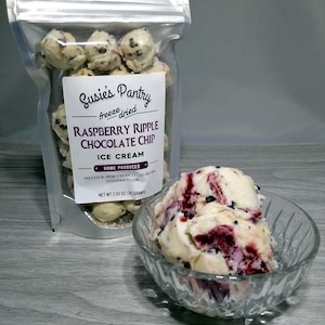 May include: A clear bag of freeze-dried Raspberry Ripple Chocolate Chip ice cream with a label that reads "Susie's Pantry". A scoop of ice cream in a glass bowl is next to the bag. The ice cream has a white base with red and dark chocolate swirls.