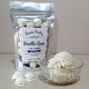 May include: A silver resealable bag filled with white freeze-dried vanilla bean ice cream bites. The bag has a label that reads "Susie's Pantry - freeze dried - Vanilla Bean - Ice Cream - Home Produced - 349 E 320 N Orem Utah 84057 | 801 800 8444 - Susiespantry.com - Net Wt 2 oz (57 grams)". A small glass bowl filled with vanilla bean ice cream is next to the bag.