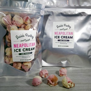 May include: A bag of freeze-dried Neapolitan ice cream. The bag is labeled "Susies Pantry" and has a pink, white, and chocolate ice cream design. The bag is open and there are several pieces of freeze-dried ice cream on a wooden surface.