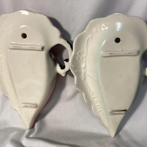 May include: Two vintage, off-white ceramic wall pockets, shaped like leaves. Each has a handle, a hole for hanging, and a small bar for support. One has red accents, the other has blue. The edges are scalloped.