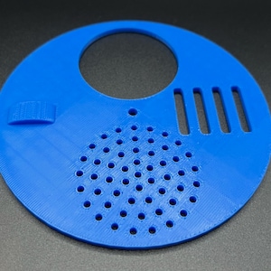 May include: A blue plastic beehive entrance reducer with a round opening, a small flap, and a grid of holes. The reducer has four vertical slots.