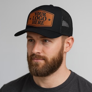 May include: Black trucker hat with a mesh back and a curved brim. The front features a brown leather patch with the text "YOUR LOGO HERE" in white. The hat is designed for a comfortable fit and is suitable for various outdoor activities.