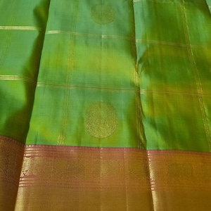 May include: Green silk saree with a gold border and intricate gold designs. The saree has a subtle ombre effect, transitioning from a darker green to a lighter green.