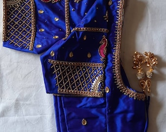 Op de afbeelding: Een blauwe zijden blouse met ingewikkelde gouden en zilveren borduurwerk. Het borduurwerk heeft een pauwenmotief en is versierd met parels en kralen.