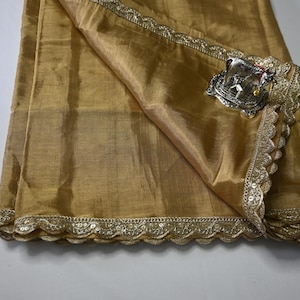 May include: A gold fabric shawl with a decorative border of sequins and a silver metal ornament. The shawl is folded in half and the ornament is on the top fold.