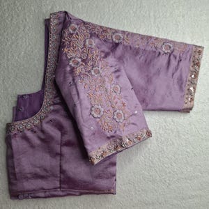 May include: A lavender-colored blouse with intricate floral embroidery on the sleeves and neckline. The sleeves and neckline are embellished with sequins. The blouse is made of a shiny, silky fabric.
