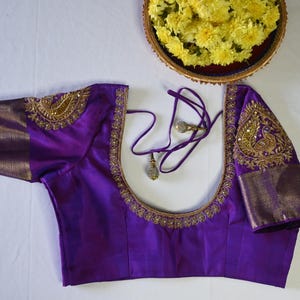 Puede incluir: Una blusa de seda morada con bordados dorados que representan un diseño de pavo real. La blusa tiene un escote redondo y mangas cortas.