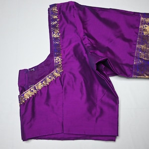 May include: A rich purple blouse with gold and blue detailing. The blouse is made from a lustrous material and has a gold pattern along the neckline and sleeve edges. The sleeves are short and the garment is neatly folded.