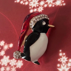 Può includere: Spilla a forma di pinguino natalizio. Il pinguino, bianco e nero, indossa un cappello da Babbo Natale rosso con una fascia e un pompon di cristalli. Lo sfondo è rosso con motivi a stella bianchi.