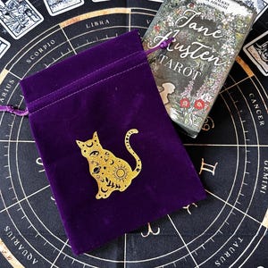 Può includere: Un sacchetto per tarocchi in velluto viola con un disegno di gatto dorato, accanto a un mazzo di tarocchi di Jane Austen. La borsa è su un tappetino nero con simboli zodiacali e immagini di carte. Il mazzo di tarocchi ha un design floreale.