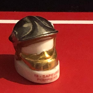 Peut inclure: Une figurine en céramique blanche représentant un pompier portant un casque argenté avec un masque facial doré. La figurine est inscrite "18 - SAPEURS POMPIERS".
