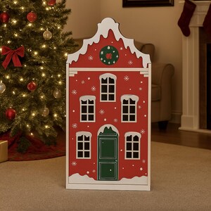 Könnte beinhalten: Rote Holz-Weihnachtsdekoration in Form eines Hauses mit weißem, schneebedecktem Dach und Fensterrahmen. Das Haus hat eine grüne Tür, schwarze Fenster und Details von Schneeflocken und Sternen. Im Hintergrund ein geschmückter Weihnachtsbaum.