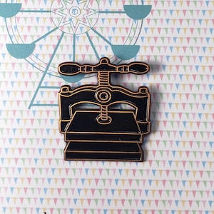 May include: A black and gold enamel pin in the shape of a printing press. The pin is on a card with a Ferris wheel design and the text "The Paper Carnival" and "thepapercarnival.com". The background has a pattern of colorful triangles.