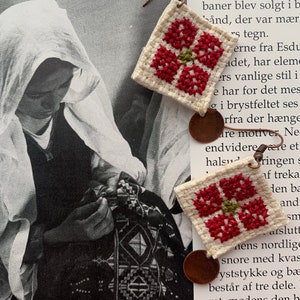 Könnte beinhalten: Ein Paar handgefertigte Ohrringe mit einer weißen Stoffbasis und roter und grüner Kreuzstich-Stickerei. Die Ohrringe sind an einem braunen Metallhaken befestigt.