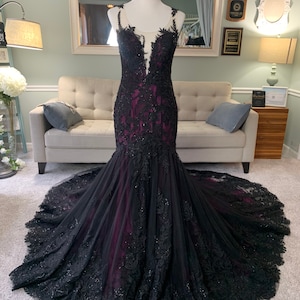 May include: A black and burgundy mermaid style gown with lace detailing and a sheer bodice. The gown has a long train and is fitted to the body.