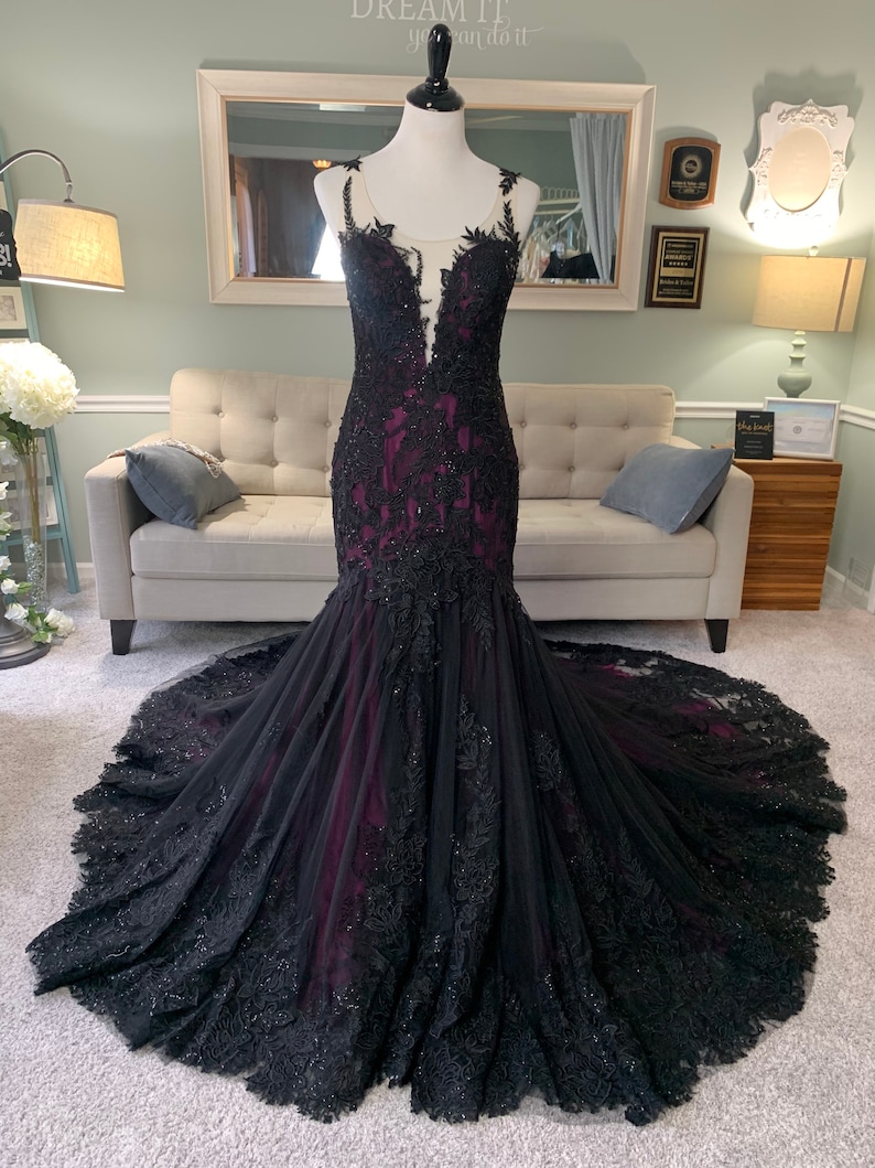 May include: A black and burgundy mermaid style gown with lace detailing and a sheer bodice. The gown has a long train and is fitted to the body.