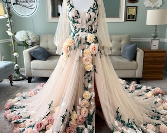 Puede incluir: Un vestido de tul rosa claro con un escote en V profundo y mangas largas. El vestido está adornado con aplicaciones florales 3D en tonos rosa, melocotón y blanco, con hojas verdes. El vestido tiene una abertura en la pierna derecha.