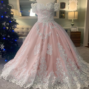 May include: A pink off-the-shoulder ball gown with white lace detailing. The dress features a fitted bodice and a full skirt, with intricate lace patterns adorning the fabric. The gown is displayed on a mannequin.
