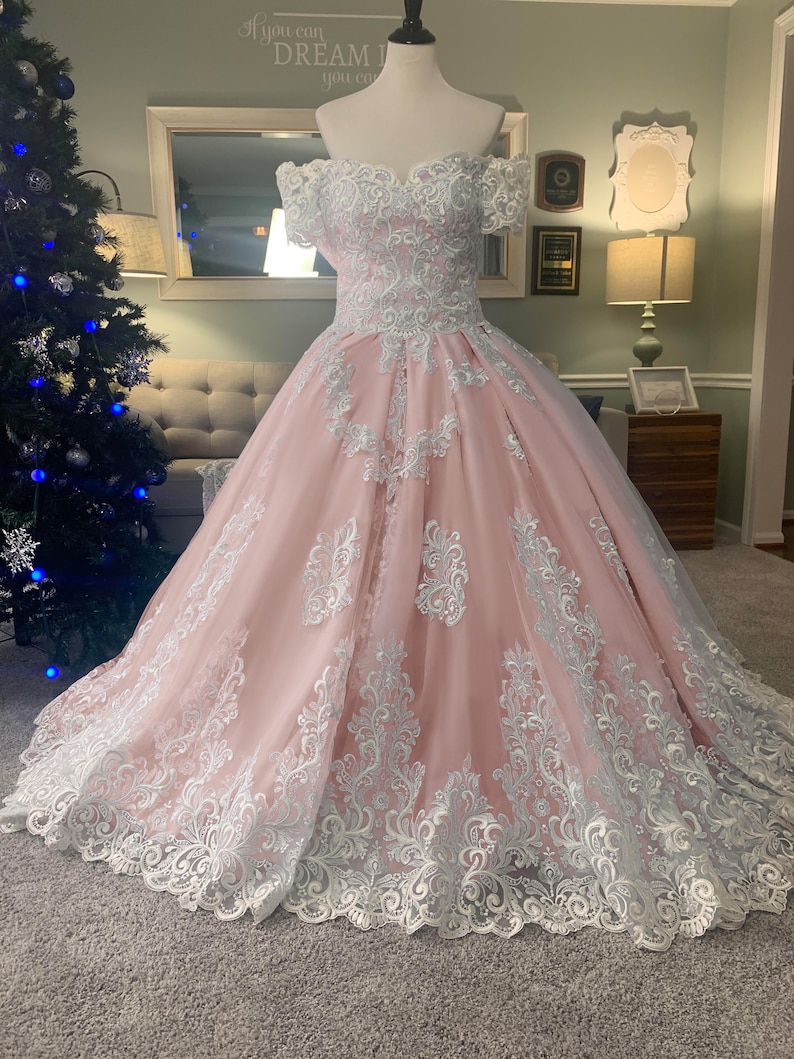 May include: A pink off-the-shoulder ball gown with white lace detailing. The dress features a fitted bodice and a full skirt, with intricate lace patterns adorning the fabric. The gown is displayed on a mannequin.