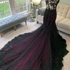 May include: A black and burgundy mermaid style gown with lace detailing and a long train. The gown has a fitted bodice and a flowing skirt.