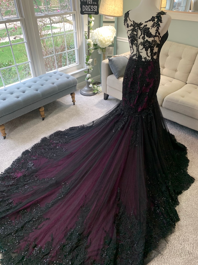May include: A black and burgundy mermaid style gown with lace detailing and a long train. The gown has a fitted bodice and a flowing skirt.