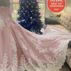 May include: A pale pink wedding dress with a long train, adorned with detailed white lace. The dress is shown near a window with a Christmas tree in the background. The Knot Best of Weddings 2020 logo is visible.