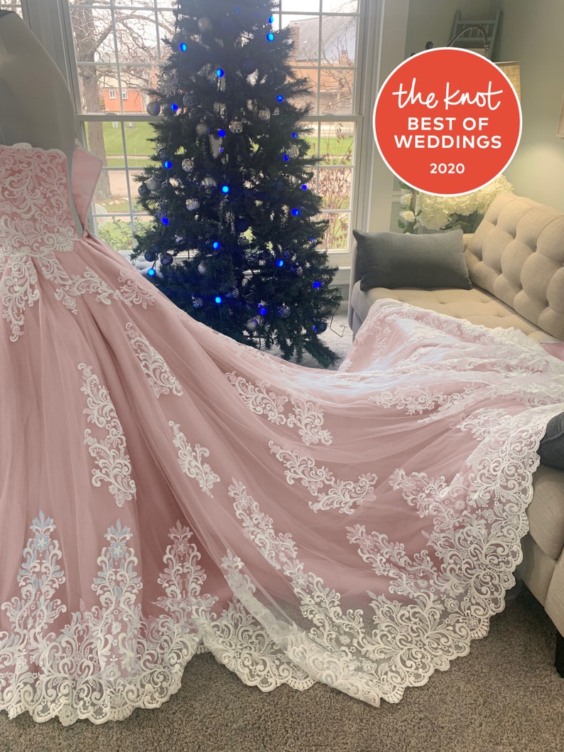 May include: A pale pink wedding dress with a long train, adorned with detailed white lace. The dress is shown near a window with a Christmas tree in the background. The Knot Best of Weddings 2020 logo is visible.