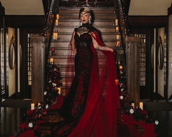 May include: A floor-length black and red gown featuring a sweetheart neckline, intricate lace detailing, and a sheer red cape that extends into a long train. The garment is fitted through the bodice and hips, flaring out at the bottom hem.