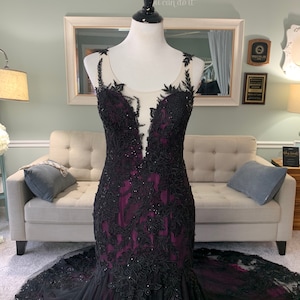 May include: A black and purple mermaid-style gown with lace detailing and a plunging neckline. The gown has a sheer bodice and a full skirt.