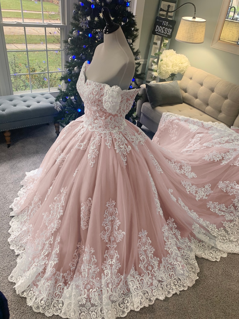 May include: A blush pink off-the-shoulder wedding dress with detailed white lace. The ballgown has a full skirt and a long train, displayed on a mannequin. The dress is decorated with floral lace patterns.