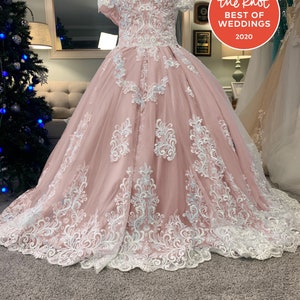May include: A blush pink off-the-shoulder wedding dress with white lace detailing. The bodice and full skirt feature intricate floral lace patterns. The dress is displayed on a mannequin, with a "Best of Weddings 2020" sticker visible.