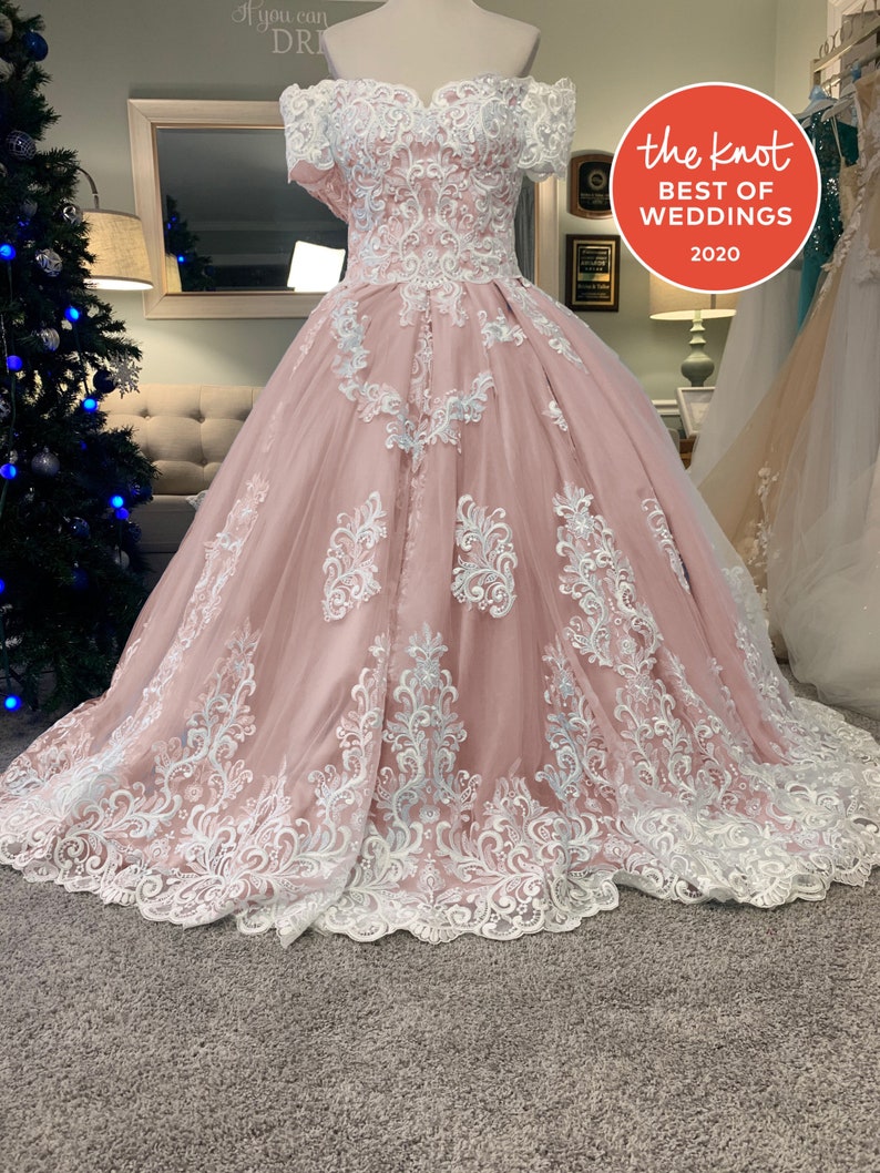 May include: A blush pink off-the-shoulder wedding dress with white lace detailing. The bodice and full skirt feature intricate floral lace patterns. The dress is displayed on a mannequin, with a "Best of Weddings 2020" sticker visible.
